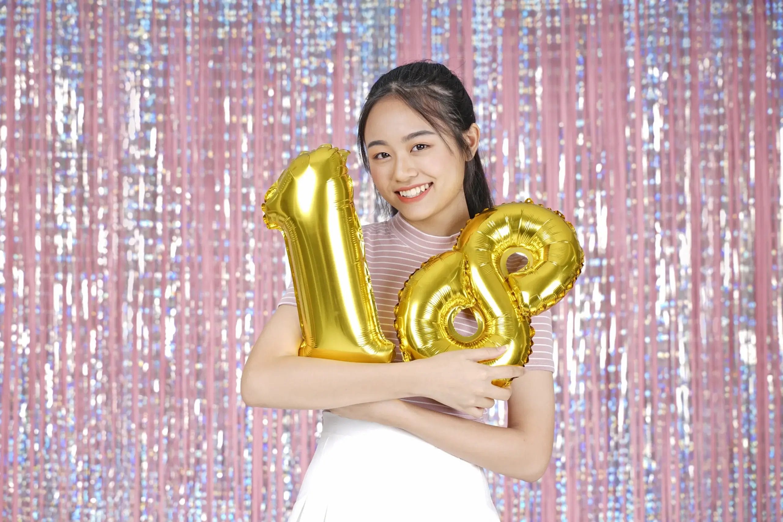A cheerful young woman with dark hair holds two gold-colored number balloons in front of a shiny pink background, celebrating a birthday. The woman wears a striped top and a white skirt, smiling brightly with her arms around the balloons. The foil background suggests a festive indoor location.